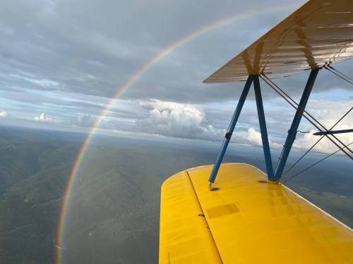 A vibrant rainbow arcs over green mountains, viewed from the wing of a yellow biplane against a cloudy sky.