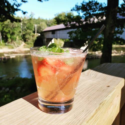 A refreshing cocktail with mint and strawberries, placed on a wooden railing by a river with trees in the background.