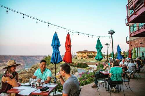 A lively outdoor dining scene with people enjoying drinks, colorful umbrellas, and a scenic view in the background.