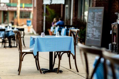 A small outdoor restaurant table with a blue checkered tablecloth and two chairs, set on a sidewalk.