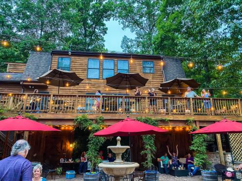 A cozy outdoor restaurant with string lights, umbrellas, and guests enjoying the ambiance on a wooden deck surrounded by trees.