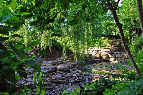 A serene garden scene featuring a stone pathway, a pond, and a green bridge surrounded by lush foliage.