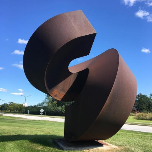 Abstract metal sculpture with curved, intertwined shapes, set against a blue sky and green grass.