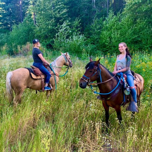 Two women riding horses in a lush green field surrounded by trees and wildflowers.