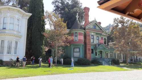 A colorful Victorian-style house surrounded by trees, with people gathered on the lawn.
