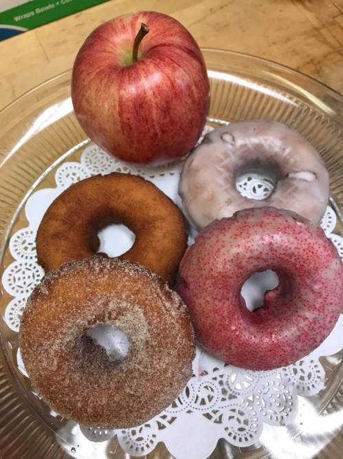A plate with four donuts—two glazed, one cinnamon sugar, and one pink sprinkle—next to a red apple.