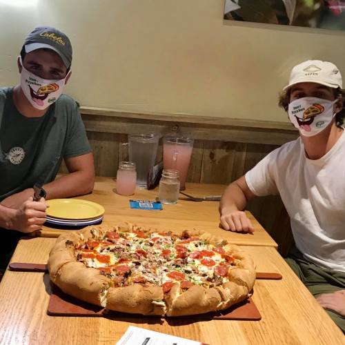 Two young men wearing masks sit at a table with a large pizza in the center, smiling at the camera.
