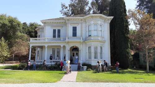 A large, white Victorian house with a porch, surrounded by trees, with a group of people gathered outside.