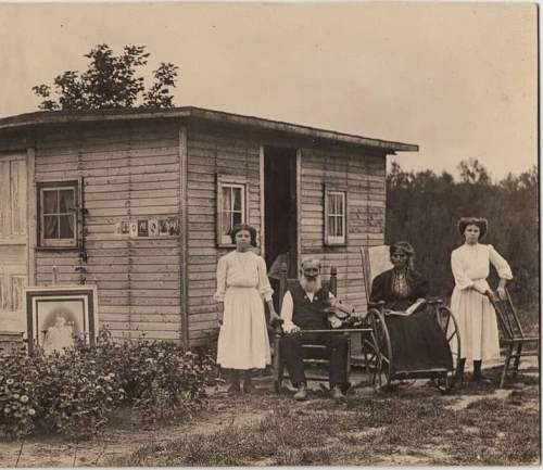 A sepia-toned photo of four people outside a small wooden house, with two women standing, a man seated, and another woman in a wheelchair.