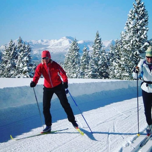 Two skiers glide on a snowy trail with mountains and evergreen trees in the background under a clear blue sky.