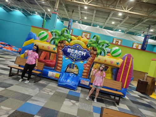 Two people in matching pink shirts stand beside a colorful inflatable beach-themed bounce house.