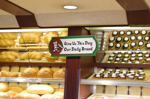 A bakery display featuring various breads and pastries, with a sign that reads "Give Us This Day Our Daily Bread."
