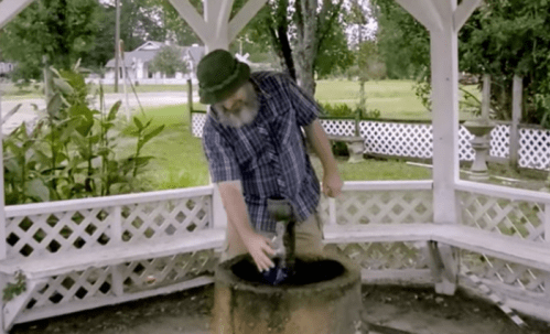 A man in a plaid shirt and hat reaches into a stone well under a gazebo surrounded by greenery.