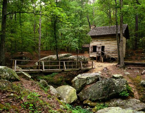 A rustic wooden cabin surrounded by trees, with a small bridge over a rocky stream in a serene forest setting.
