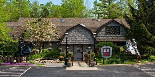 A charming lodge with a shingled roof, surrounded by greenery and flowers, featuring knight statues on horseback at the entrance.