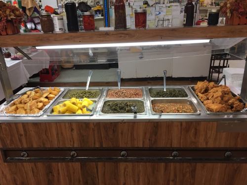 A buffet display featuring fried chicken, greens, corn, rice, and pastries in metal trays on a wooden counter.