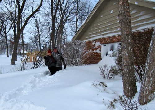 Two people stand in deep snow near a house, surrounded by trees, with snow covering the ground and building.