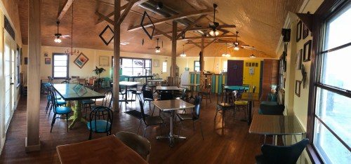 Bright, cozy café interior with colorful tables and chairs, wooden beams, and large windows letting in natural light.