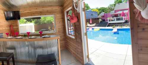 View from a cozy wooden bar area overlooking a pool with colorful lounge chairs and a vibrant backyard setting.