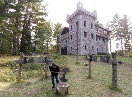 A person chops wood in front of a stone castle surrounded by trees in a grassy area.