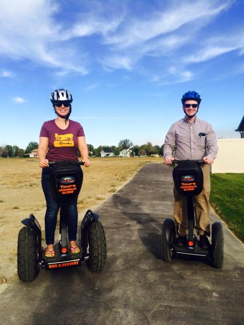 Two people riding Segways on a paved path, wearing helmets, with a clear blue sky and open fields in the background.