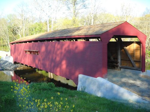 A red covered bridge spans a small stream, surrounded by greenery and wildflowers under a clear blue sky.
