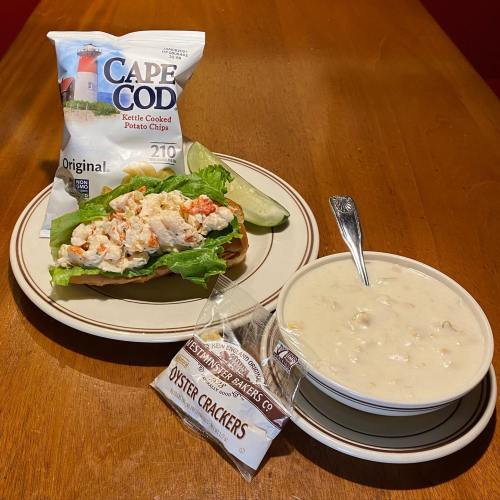 A plate with a lobster roll on lettuce, a bowl of clam chowder, a bag of chips, and a pack of oyster crackers.