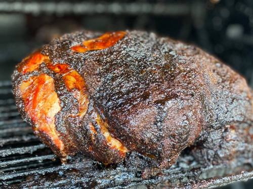 A close-up of a smoked, seasoned meat roast on a grill, showcasing a dark, crispy exterior.