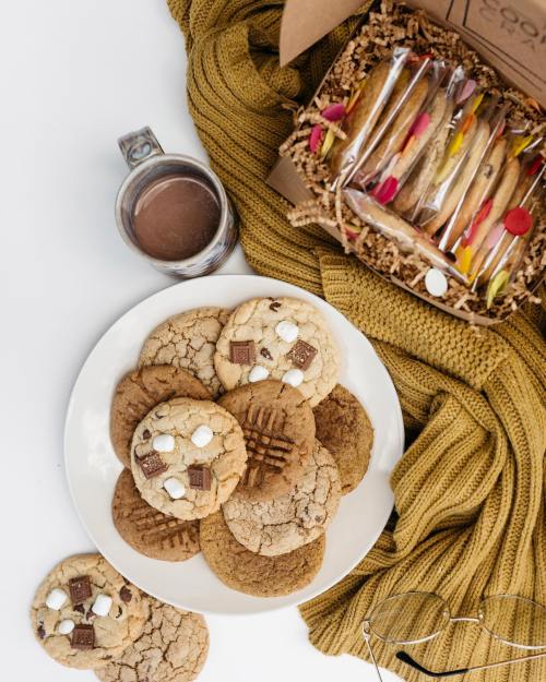 A plate of assorted cookies, a mug of hot chocolate, and a gift box of cookies on a cozy yellow knit blanket.