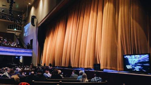 Audience seated in a theater, facing a large golden curtain and a screen displaying "Opry."