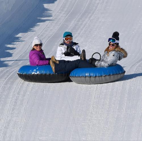 Three people in winter gear sit on inflatable tubes, ready to slide down a snowy slope. Bright sunlight illuminates the scene.