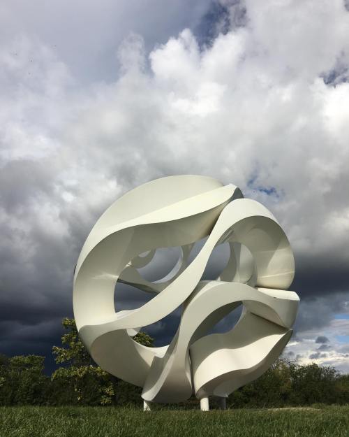 A large, abstract white sculpture with flowing curves, set against a dramatic sky with dark clouds.