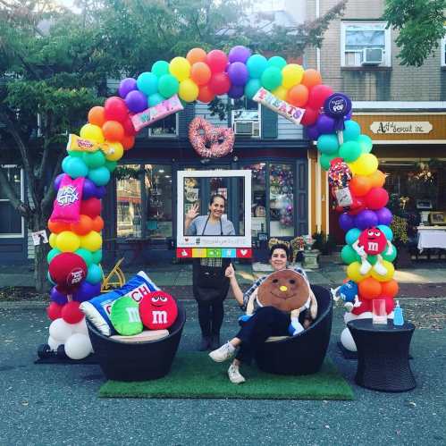 A colorful balloon arch with treats-themed decorations, featuring two people sitting on chairs in a festive outdoor setting.