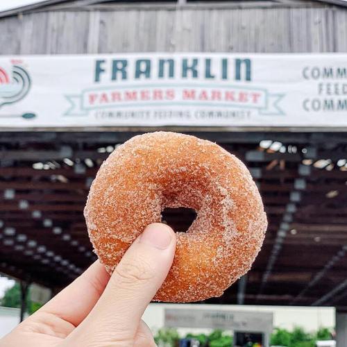A hand holds a sugar-coated donut in front of the Franklin Farmers Market sign.