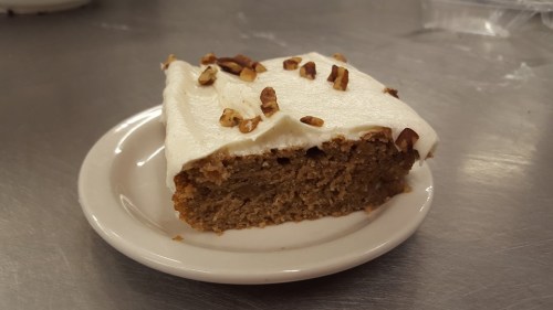 A slice of cake with cream frosting and chopped nuts on top, served on a small white plate.