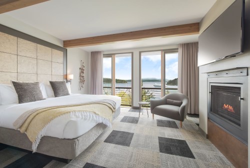 A modern hotel room with a king bed, large windows, a fireplace, and a view of a lake and mountains.