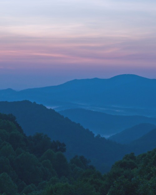 A serene mountain landscape at dusk, with layered hills and a soft purple sky.