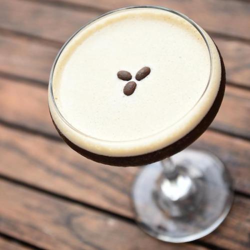 A close-up of a creamy coffee cocktail topped with three coffee beans, served in a stylish glass on a wooden table.