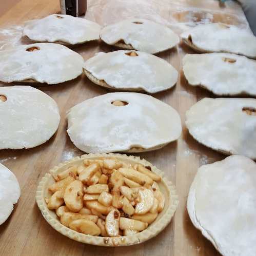 A wooden table with several dough rounds dusted with flour and a bowl of sliced, cooked apples in the foreground.