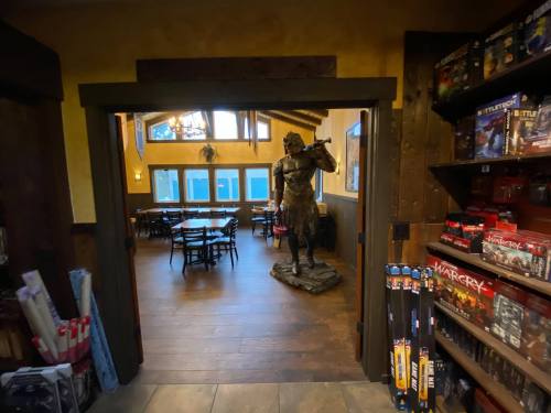 A view of a cozy room with a knight statue and tables, framed by wooden doorways and shelves of games.