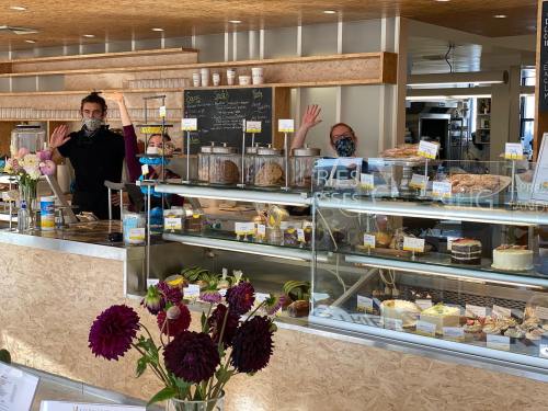 Three masked staff members wave from behind a bakery counter filled with various baked goods and flowers in the foreground.