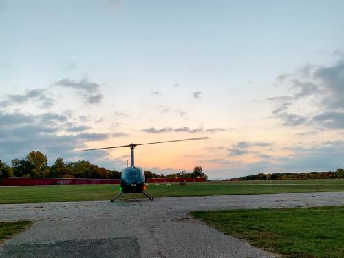 A helicopter sits on a runway at sunset, with a colorful sky and trees in the background.