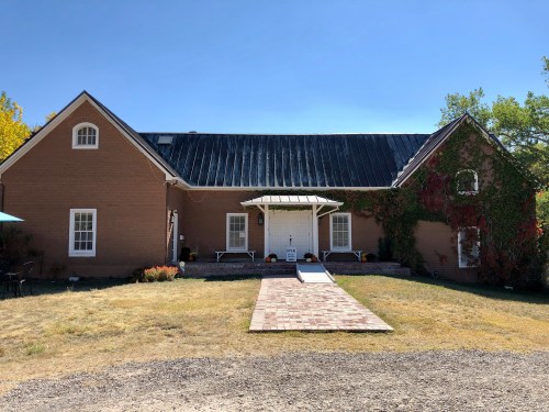 A brown, two-story building with a black roof, surrounded by grass and trees, featuring a front porch and pathway.