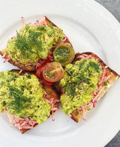 Three slices of toast topped with avocado, radish, cherry tomatoes, and fresh herbs on a white plate.