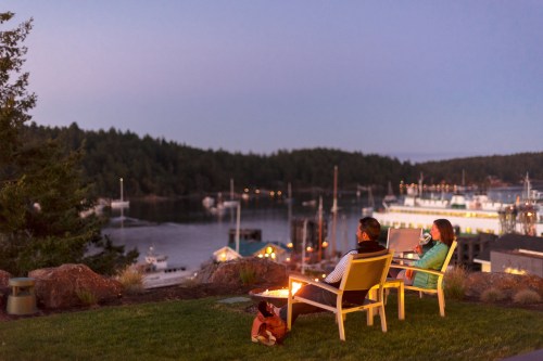 A couple sits by a fire pit, enjoying a scenic view of a harbor at dusk, surrounded by boats and trees.