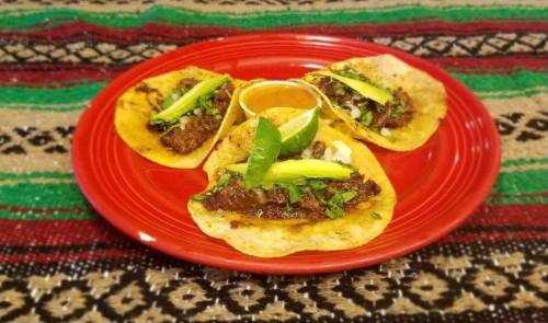 Three tacos on a red plate, topped with meat, avocado, cilantro, and lime, served with a small bowl of sauce.