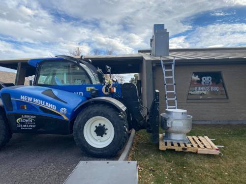 A blue New Holland telehandler parked next to a BBQ restaurant, with a ladder and equipment on a wooden pallet.