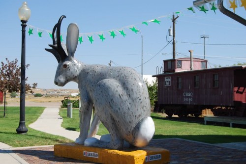 A large, gray jackrabbit statue with antlers sits on a pedestal in a park, with a red train car and green decorations in the background.
