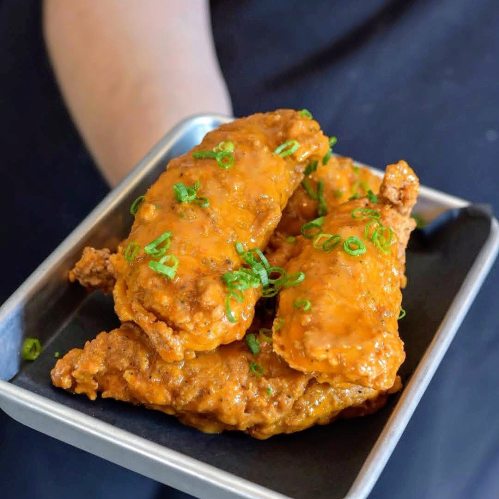 A hand holding a tray with three crispy chicken wings, coated in a spicy orange sauce and garnished with green onions.