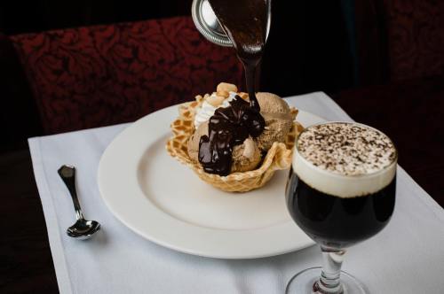 A waffle bowl filled with ice cream topped with chocolate sauce, alongside a glass of coffee with whipped cream.